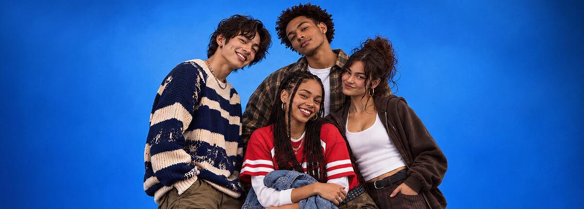 Four young adults pose together, smiling and wearing casual layered clothing against a bright blue background.