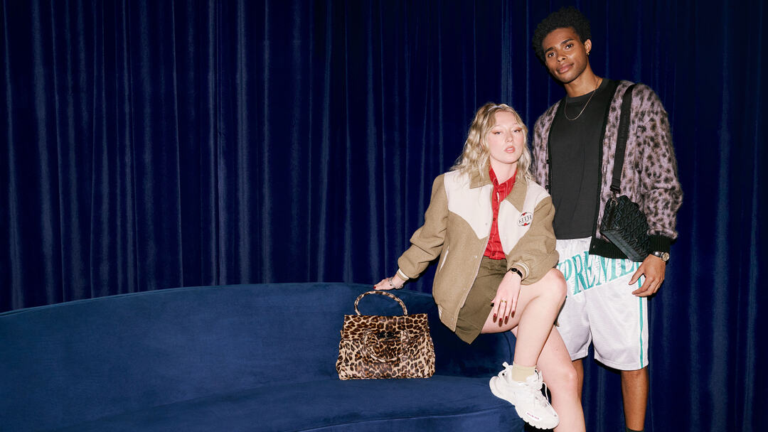 Two models wearing contemporary fashion and accessories posed beside a sofa.
