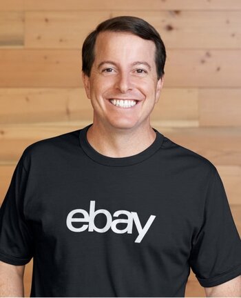 Jamie Iannone, CEO of eBay, smiles at the camera wearing a black eBay T-shirt against a light wood-paneled background.