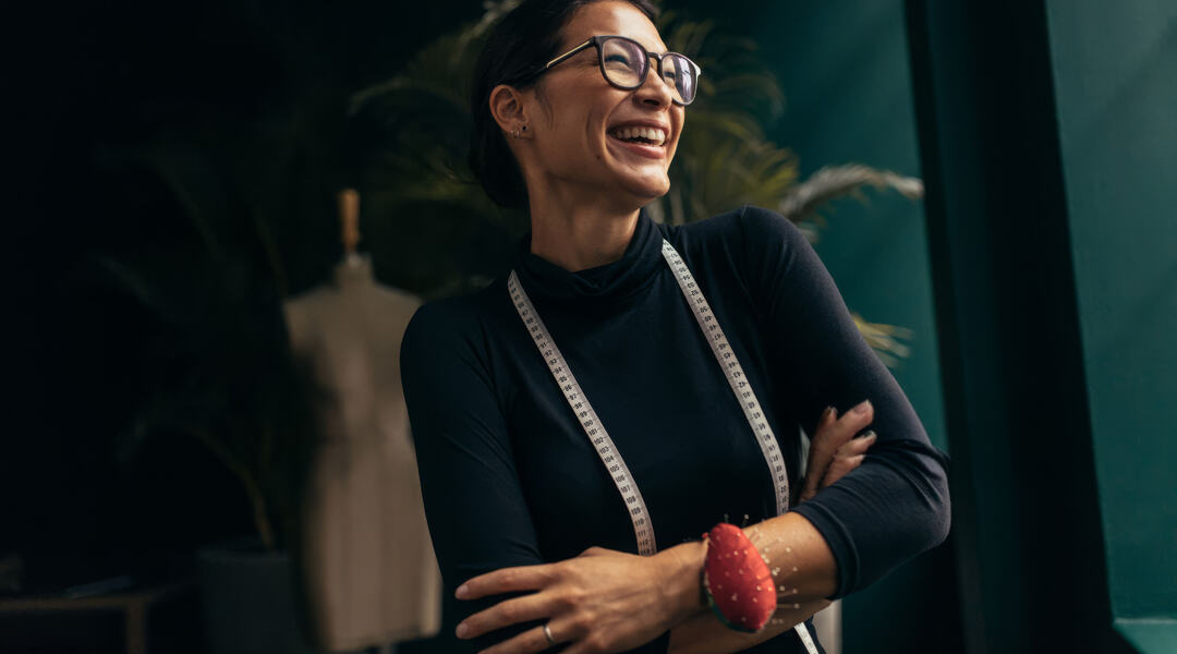 Smiling woman wearing glasses and a measuring tape around her neck stands with her arms crossed in a clothing workspace.