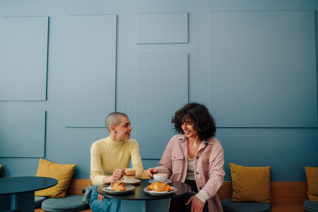 Two people sit at a café table, smiling over coffee and pastries against a blue paneled wall.