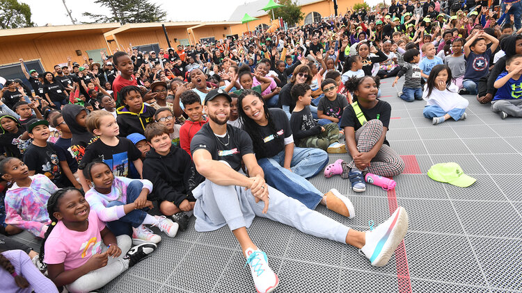 Stephen and Ayesha Curry seated and smiling alongside a big group of children.
