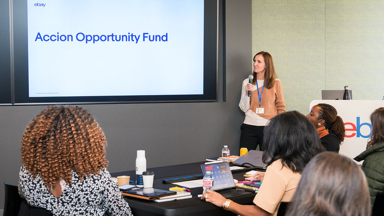 A woman stands next to a large screen displaying "Accion Opportunity Fund" while five seated attendees listen and watch during a presentation in an office setting.