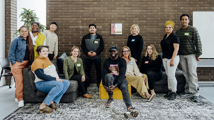 A group of thirteen people varying in gender and race posing together in a room with a brown brick wall, carpeted floor, and a whiteboard in the background. Some are standing, while others are seated on furniture.