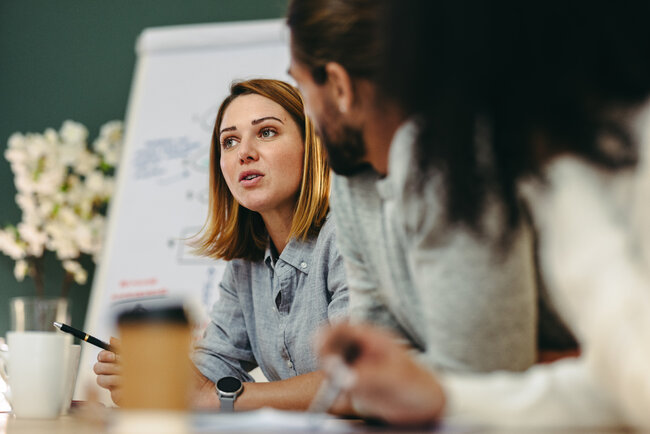 A woman speaks with colleagues during a meeting, seated at a table with a presentation board in the background.