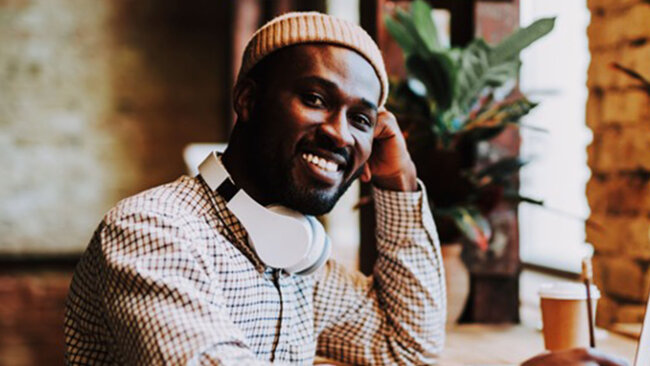 Employee smiling while working remotely with headphones around their neck in a casual workspace.