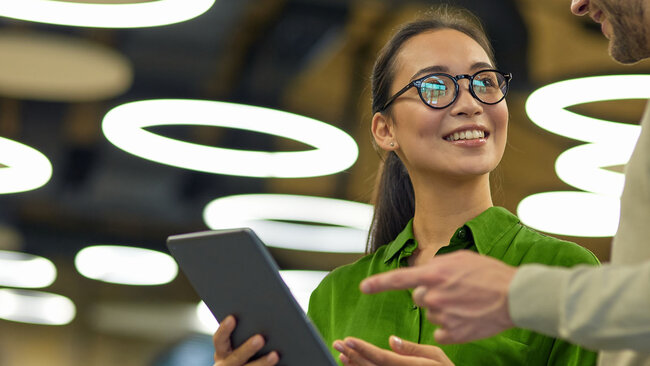 Employee holding a tablet while speaking with a colleague in an office setting.