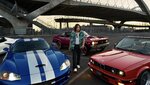 Actor and automotive enthusiast Sung Kang stands in front of four cars beneath a highway overpass: a white 2022 Chevrolet Corvette C8, blue 2006 Dodge Viper, red 1992 BMW 325i Convertible and burgundy 1975 Chevrolet Nova.