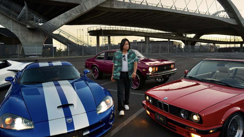 Actor and automotive enthusiast Sung Kang stands in front of four cars beneath a highway overpass: a white 2022 Chevrolet Corvette C8, blue 2006 Dodge Viper, red 1992 BMW 325i Convertible and burgundy 1975 Chevrolet Nova.